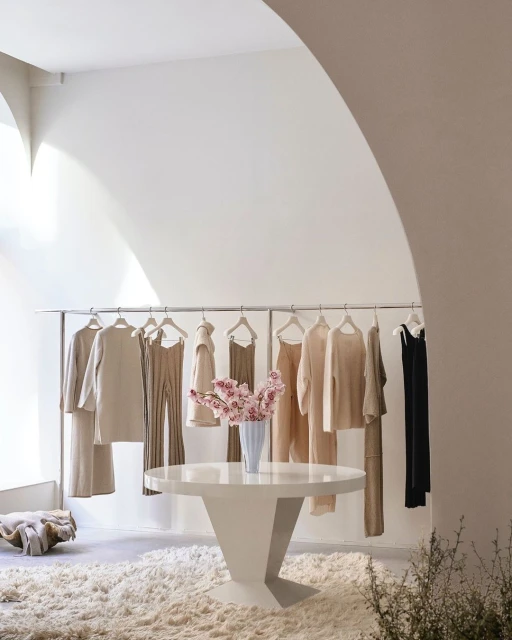 Clothing boutique interior with a round white table holding a vase of pink flowers, a rack of clothes, and a shag carpet.