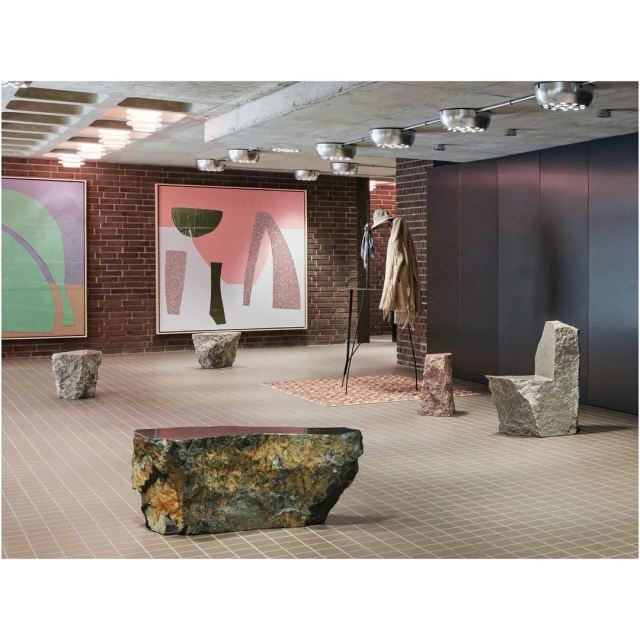 Interior showroom with stone furniture and abstract paintings on brick walls, illuminated by modern ceiling lights.