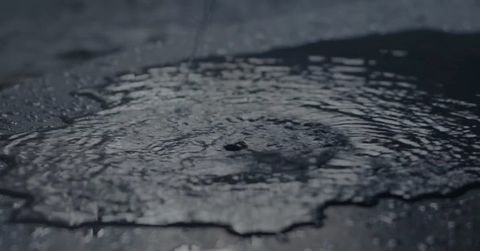 Close-up of water on dark pavement, creating ripples and reflections in natural light. The scene has a dark, wet, and moody aesthetic.