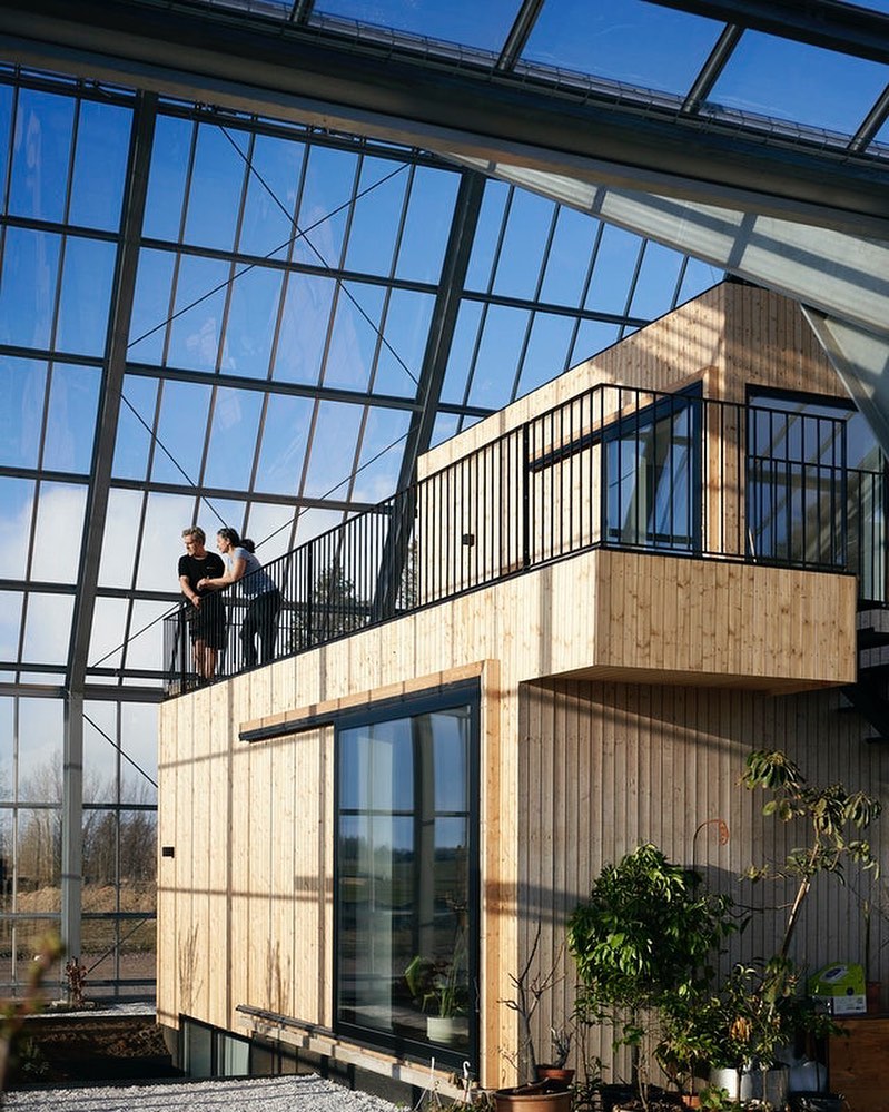 Two women stand on the second-story balcony of a modern wooden structure inside a soaring glass-roofed conservatory or greenhouse.