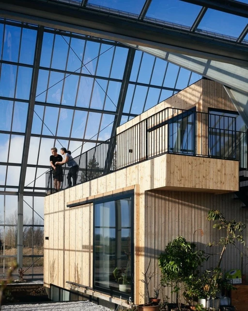 Two women stand on the second-story balcony of a modern wooden structure inside a soaring glass-roofed conservatory or greenhouse.