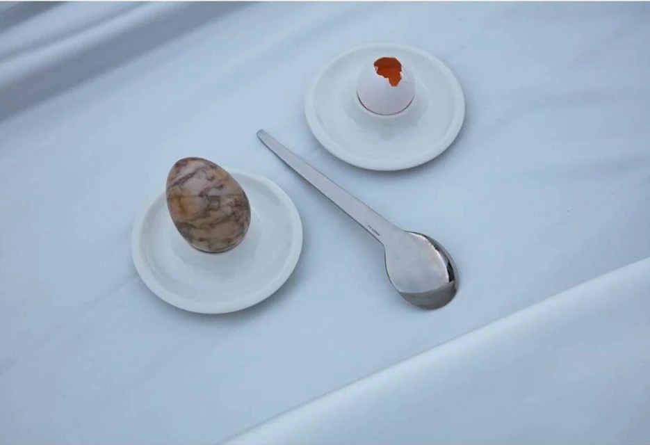 Still life photograph of a brown egg and a cracked white egg shell with a silver spoon on a white plate and white fabric.