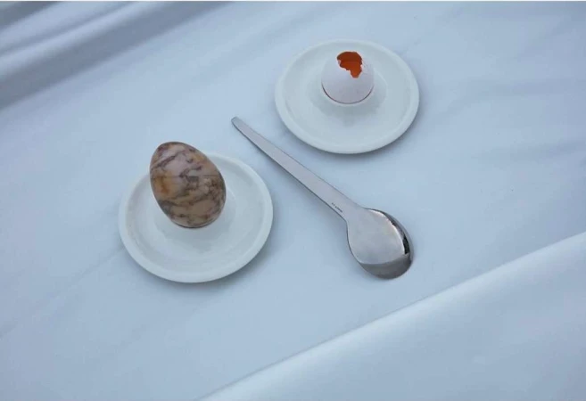 Still life photograph of a brown egg and a cracked white egg shell with a silver spoon on a white plate and white fabric.