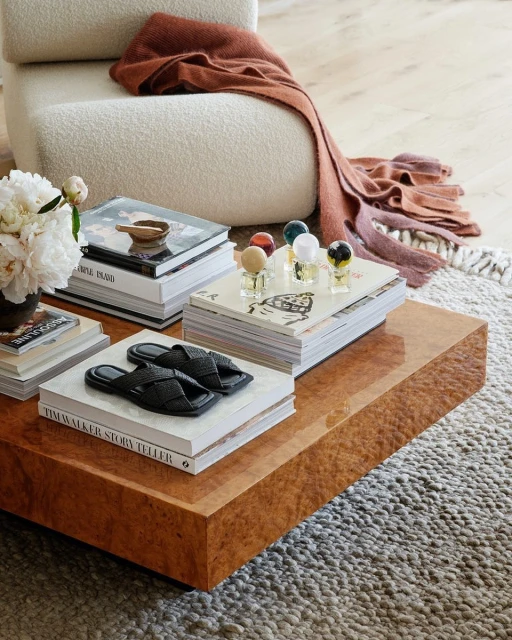 Indoor scene features a coffee table with stacked books, slippers, decorative glass orbs, a vase of flowers, and a cozy throw blanket.