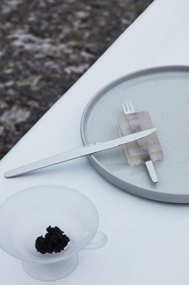 Overhead shot of a plate with a knife, fork, and unidentified food, and a bowl of black caviar on a white table.