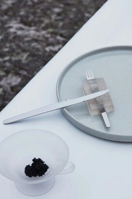 Overhead shot of a plate with a knife, fork, and unidentified food, and a bowl of black caviar on a white table.