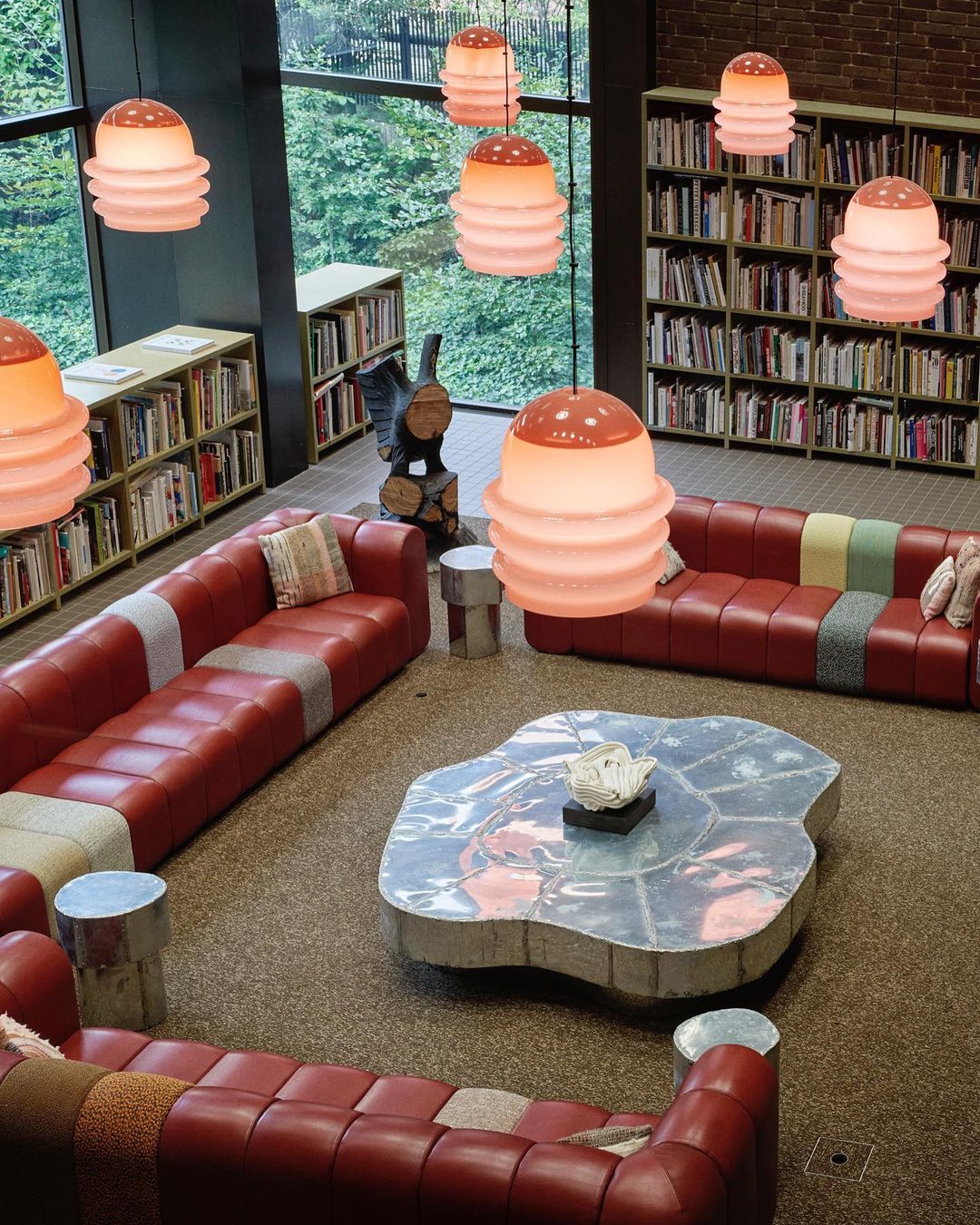 Library interior featuring red sectional sofas, a custom metal coffee table, bookcases filled with books, and modern pendant lighting.