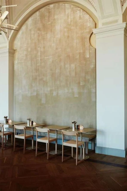 Interior view of restaurant seating area with light wood chairs and tables. Sunlight shines on the wall, casting a warm glow.