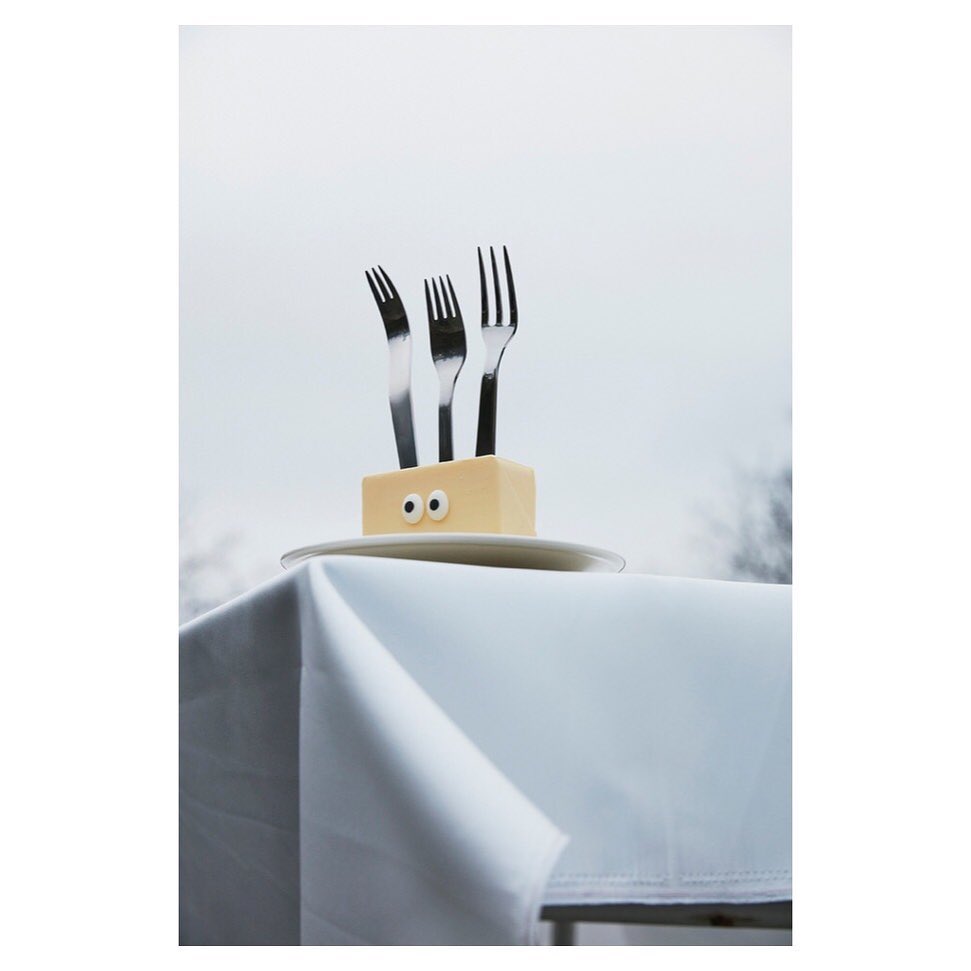 Three stainless steel forks sticking out of a butter block with googly eyes on a white plate on a white tablecloth, against sky.
