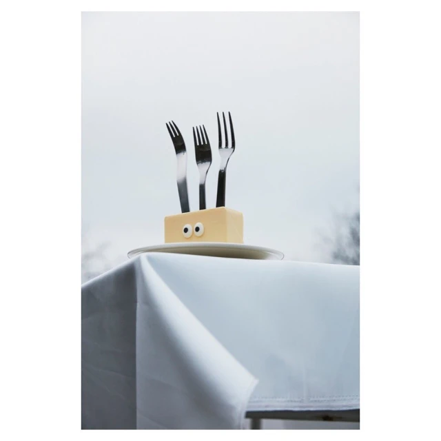 Three stainless steel forks sticking out of a butter block with googly eyes on a white plate on a white tablecloth, against sky.
