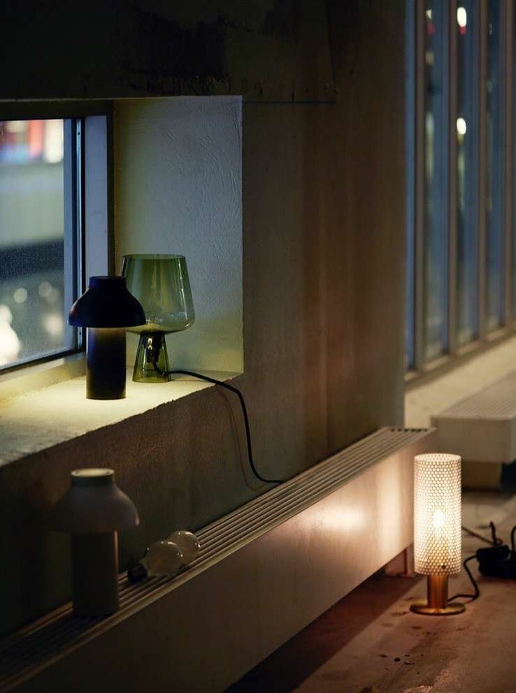 Several modern lamps of varying designs and heights are illuminated, sitting on a concrete ledge inside a dimly lit room with windows.