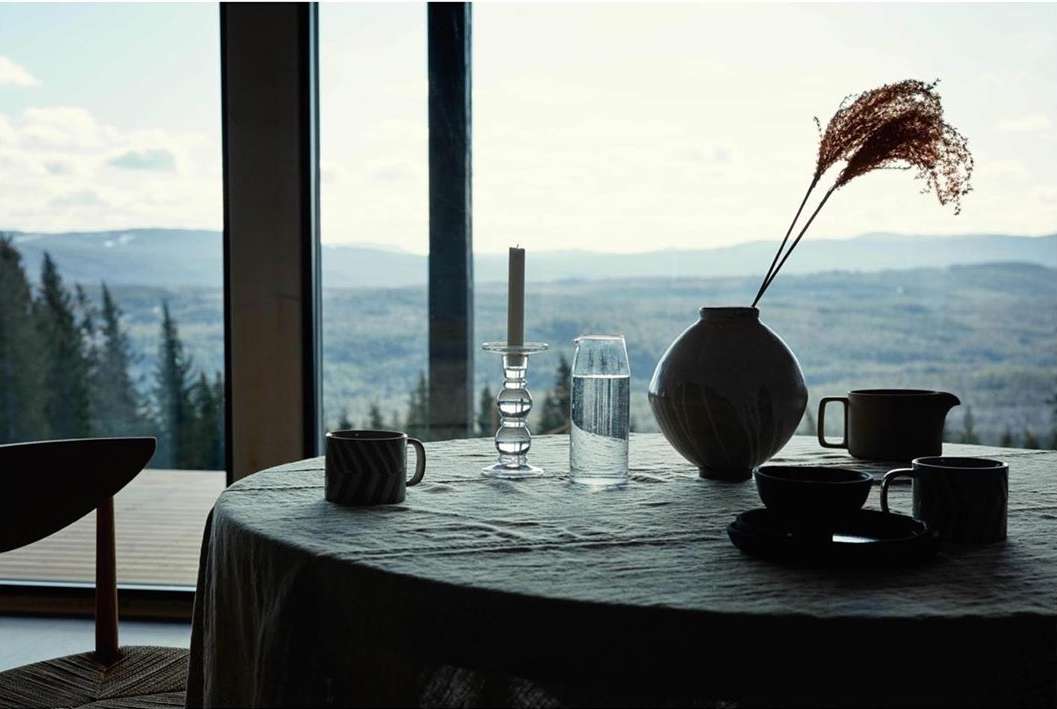 Still life of table setting with mugs, plates, vases, water pitcher, and candlesticks in front of a window with a mountain view.