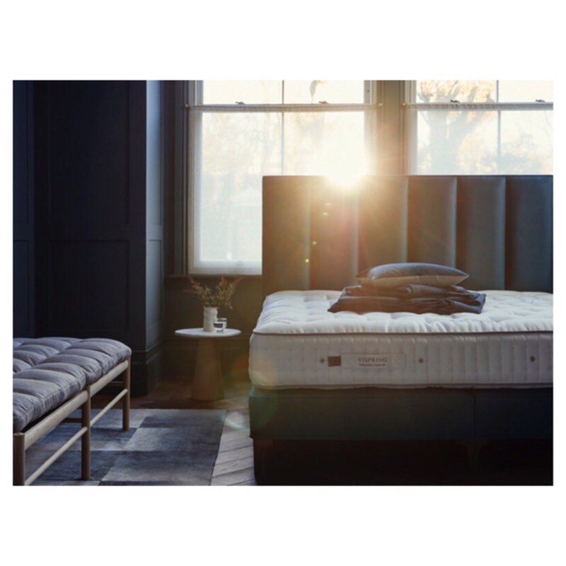 Bedroom setup with a queen-sized bed with a gray headboard, layered blankets and pillows, a bench, a table with flowers, and natural light.