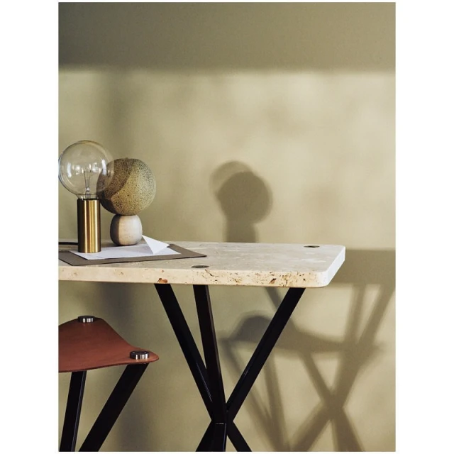 Modern interior scene featuring a table with black frame, a glass and brass lamp, spherical decor, a stool and a neutral wall.