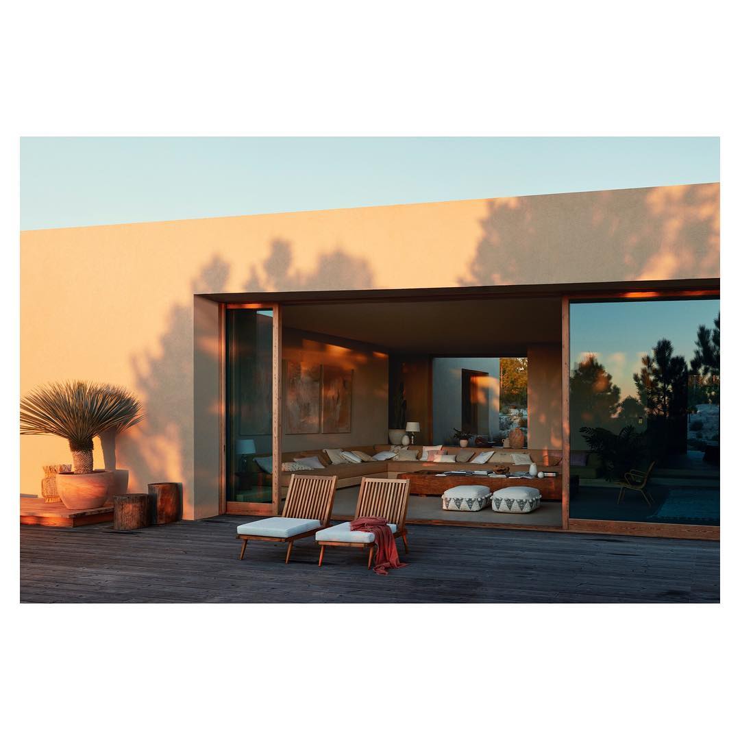 Outdoor patio featuring a pair of wooden lounge chairs with white cushions on a wooden deck, facing an open-plan living space inside a house.