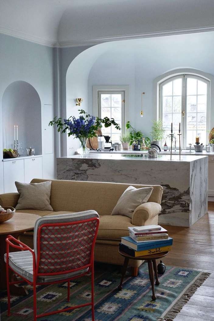 Living room interior with a tan sofa, patterned rug, red chair, marble kitchen island, hardwood floors, and natural light.