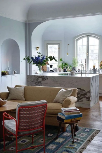 Living room interior with a tan sofa, patterned rug, red chair, marble kitchen island, hardwood floors, and natural light.