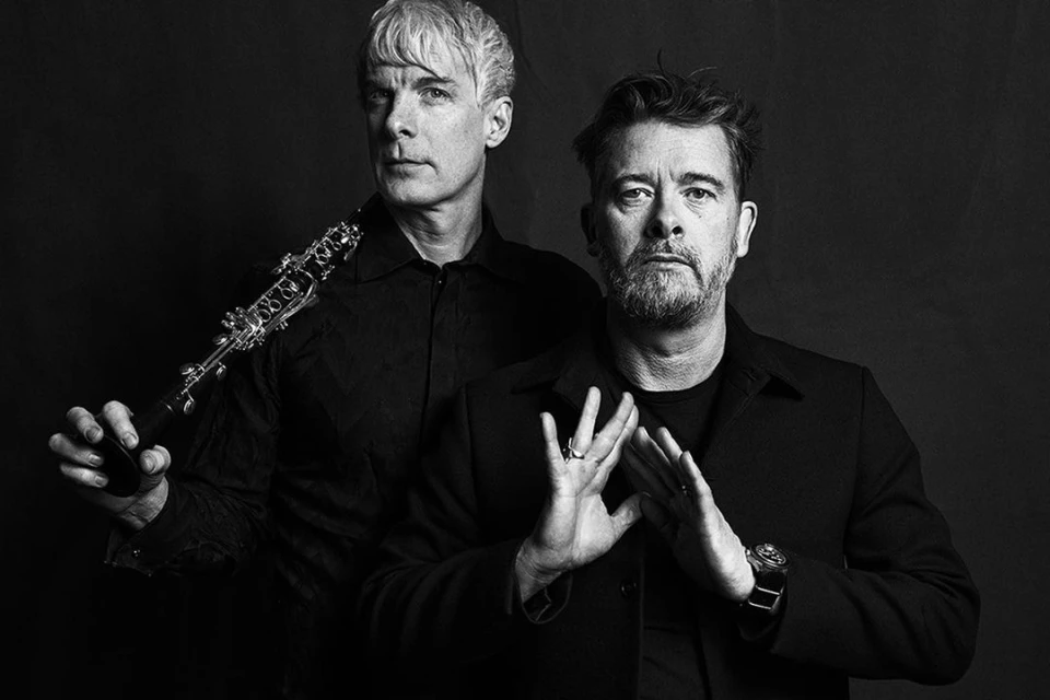 Monochrome portrait of two adult men, one holding a clarinet, posing in front of a dark background in a photo studio setting.