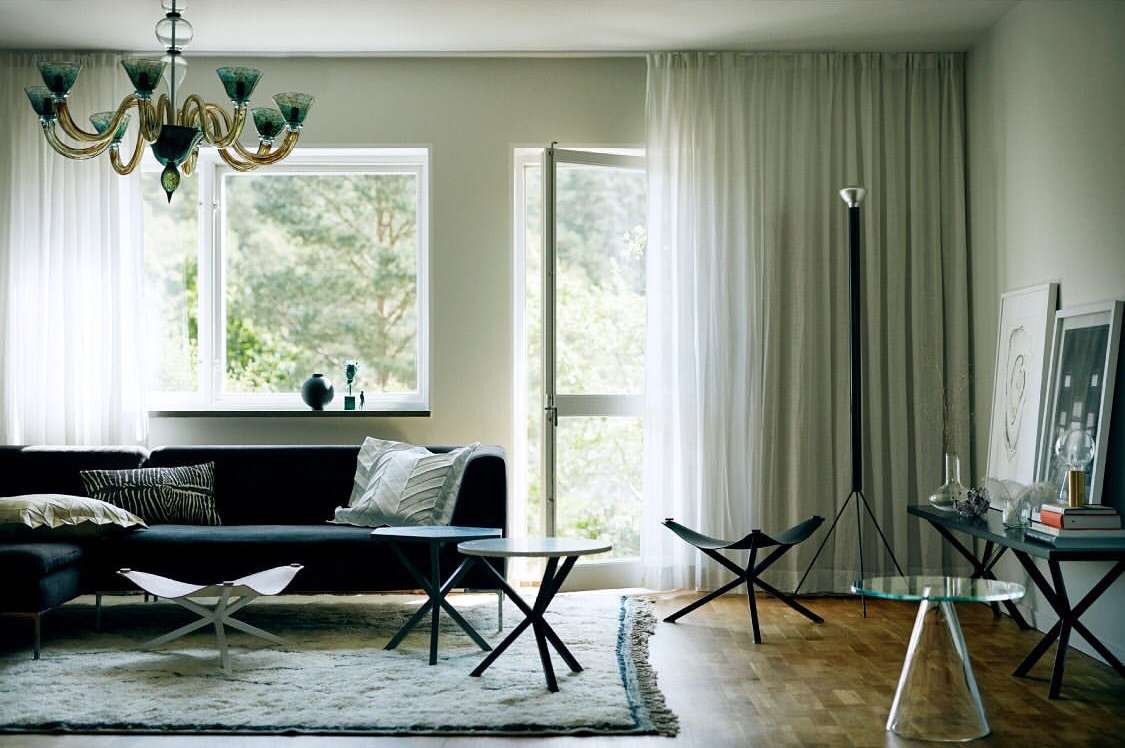 Modern living room interior with a black sofa, patterned pillows, decorative lighting fixture, and a large window offering an outdoor view.