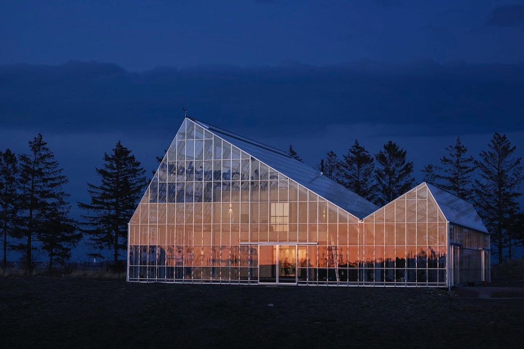 Glass building with a triangular roof at dusk, surrounded by pine trees, reflecting the orange sunset sky.