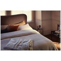 A made bed with soft grey linens, layered pillows, and a knitted throw blanket inside a room with morning sunlight.