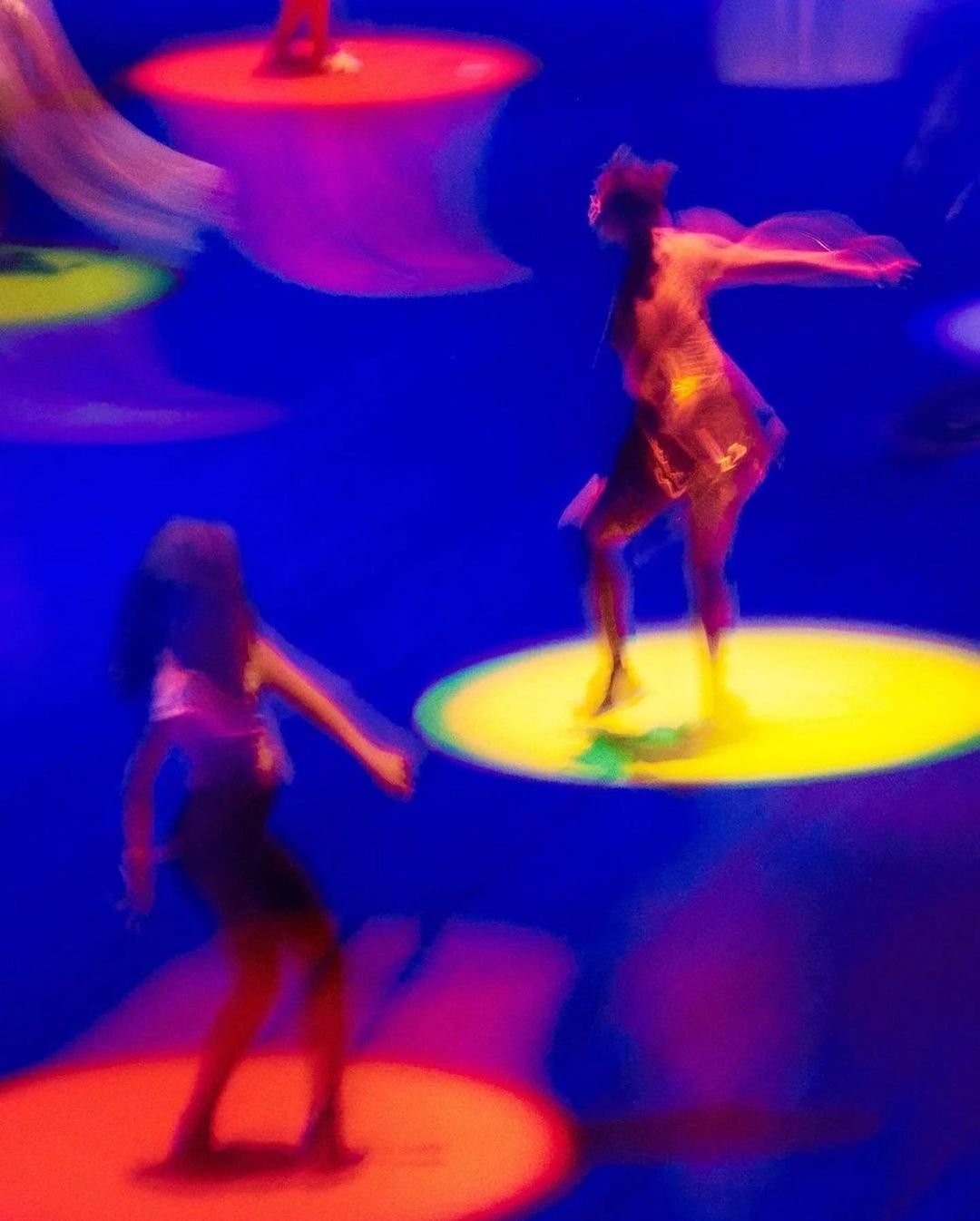 Two dancers perform on illuminated platforms in a dynamic, blurred shot. The vibrant colors and long exposure create a sense of movement.