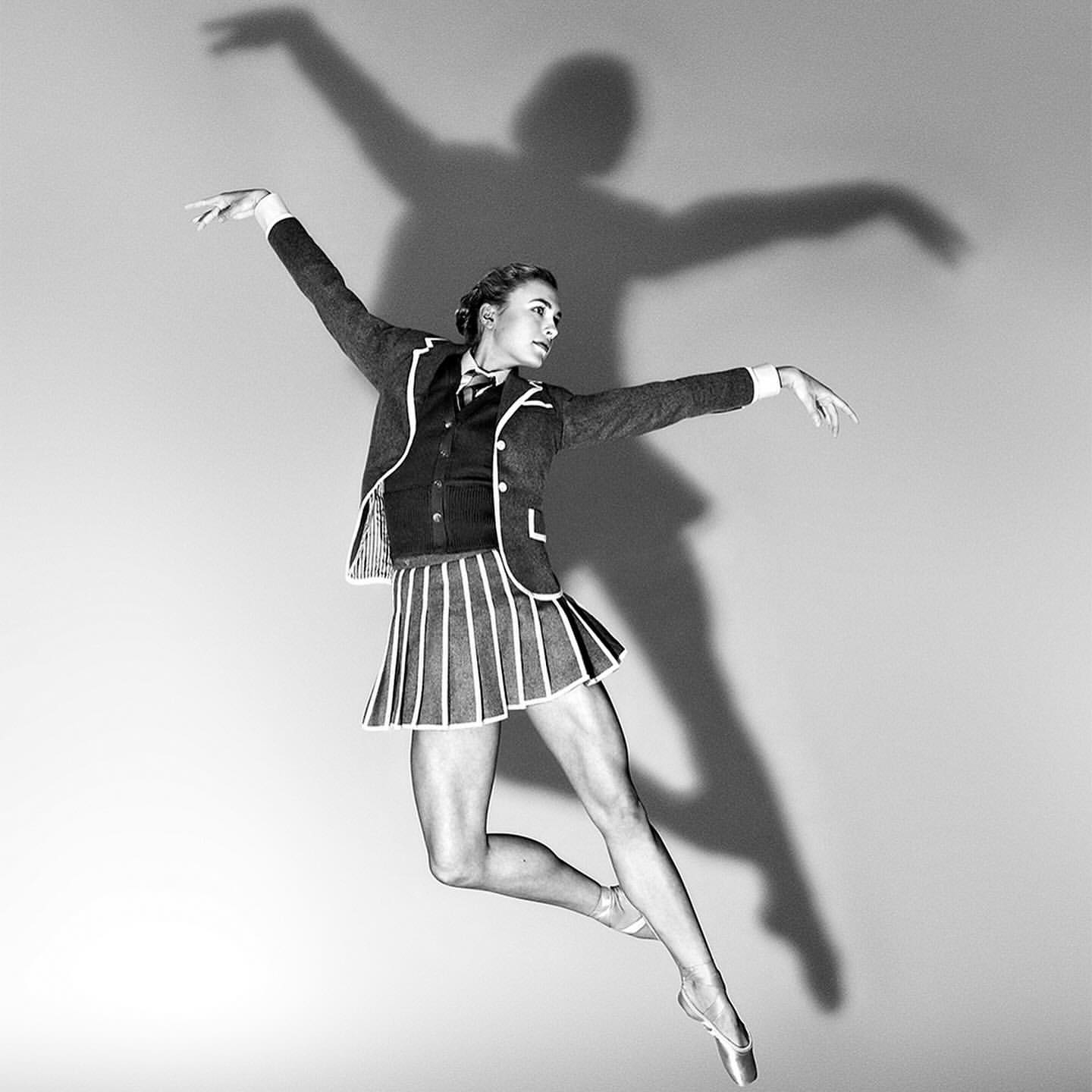 Black and white photo of a woman in a school uniform leaping, with a shadow behind her on a plain background, showcasing her athletic ballet pose.