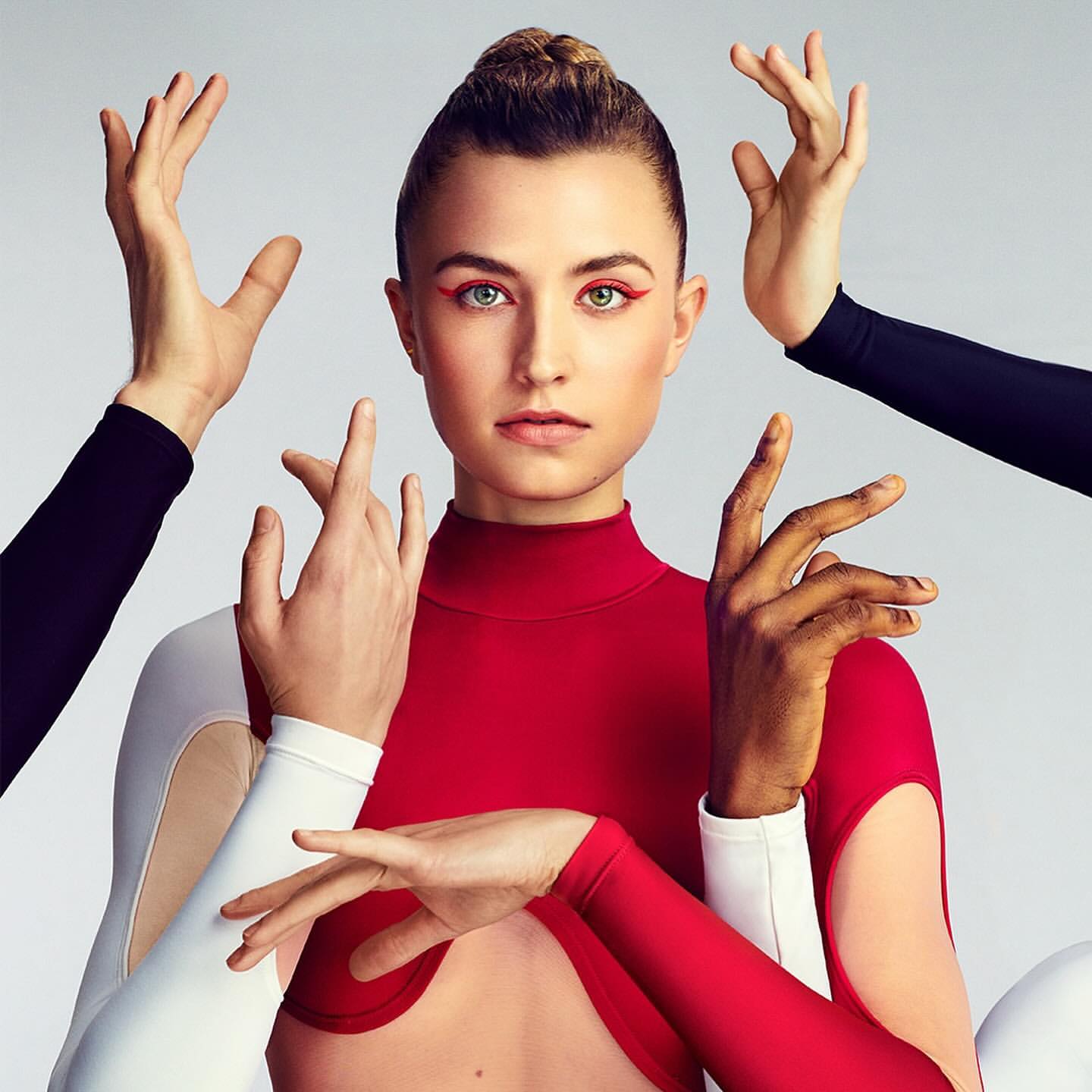 Portrait of a woman in a red top surrounded by hands against a gray background. Red eyeliner accentuates the model's green eyes.