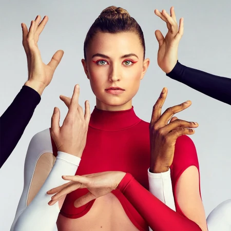 Portrait of a woman in a red top surrounded by hands against a gray background. Red eyeliner accentuates the model's green eyes.
