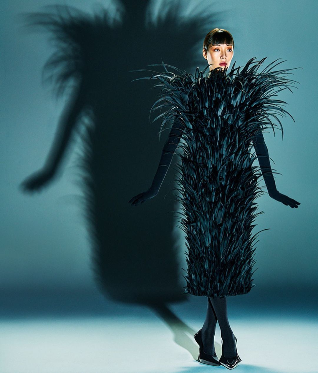 A woman in a feathered dress poses in a studio with a shadow. The dress is knee-length and made entirely of long, glossy black feathers.