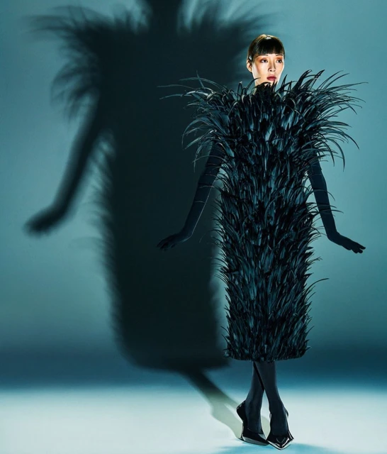A woman in a feathered dress poses in a studio with a shadow. The dress is knee-length and made entirely of long, glossy black feathers.