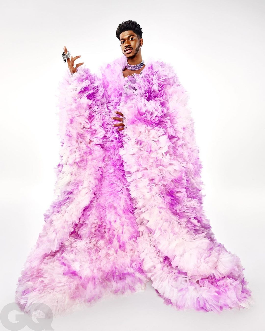 Lil Nas X poses in a voluminous lavender and white ruffled robe, wearing jewelry against a stark white backdrop in a studio setting.
