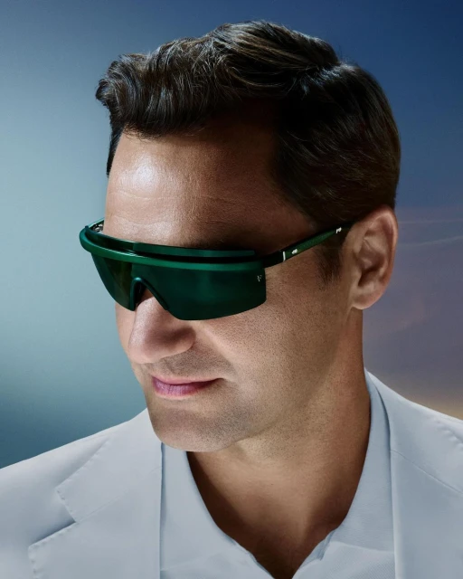 A man with dark hair and olive skin is wearing futuristic green sunglasses and a white suit jacket, looking slightly off-center.