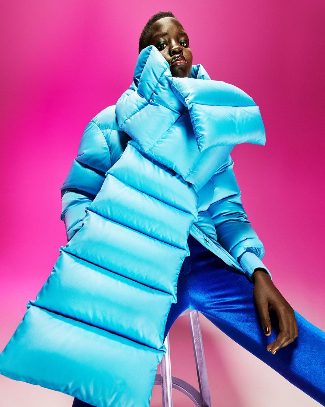 Editorial fashion shot of a Black female model in a large, light-blue puffer coat and royal-blue pants against a vibrant pink background.