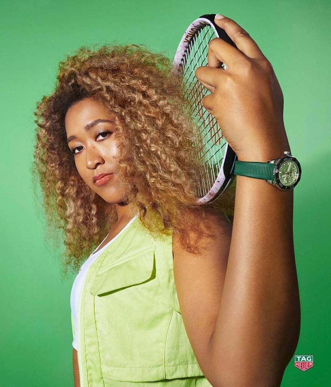Portrait of Naomi Osaka holding a tennis racket. She wears a lime green vest and a Tag Heuer watch against a green background.