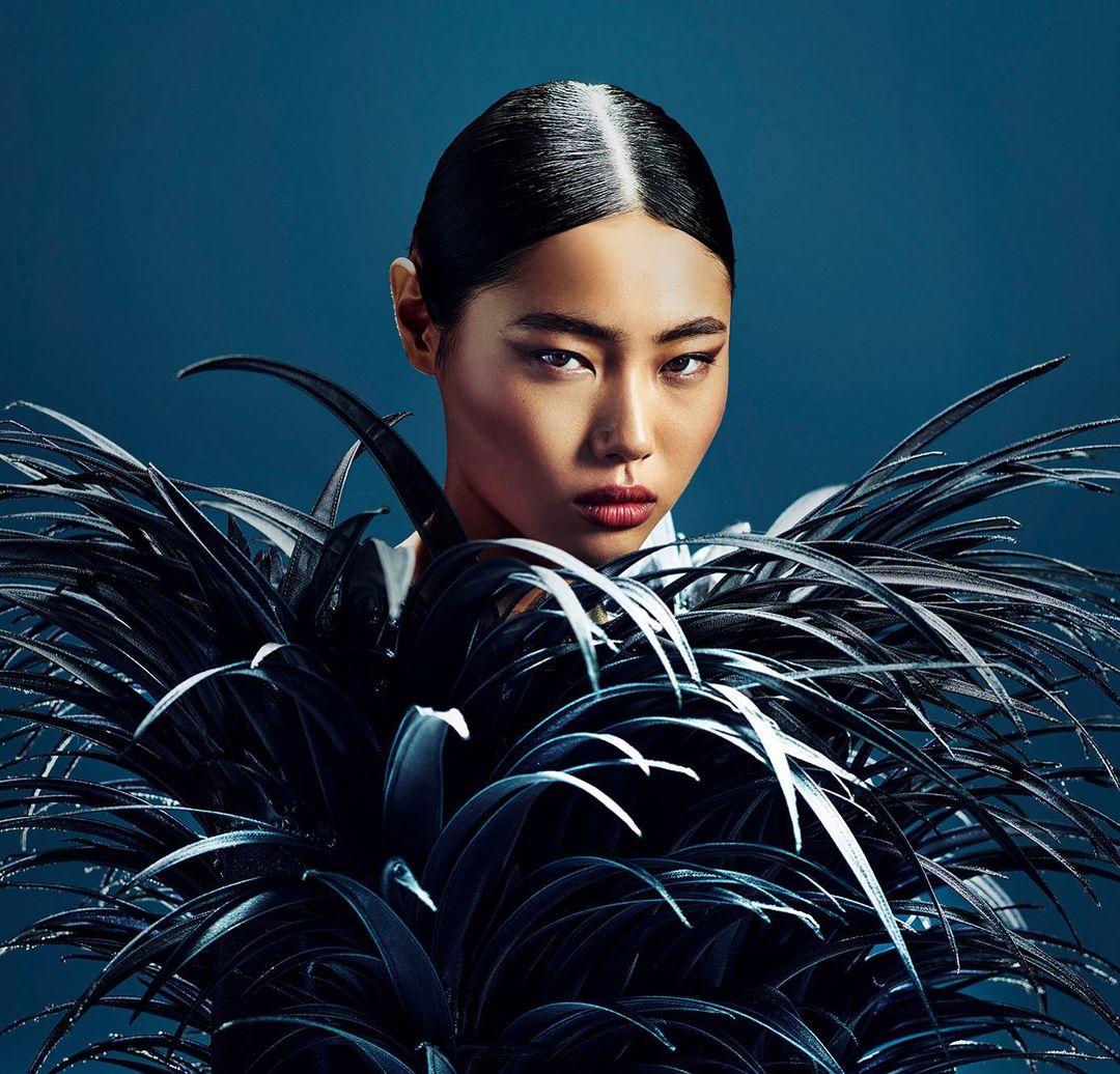 Portrait of a woman with dark hair and red lips, wearing a dark feathered garment against a dark blue backdrop in a studio setting.