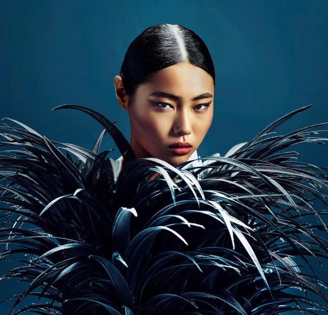 Portrait of a woman with dark hair and red lips, wearing a dark feathered garment against a dark blue backdrop in a studio setting.