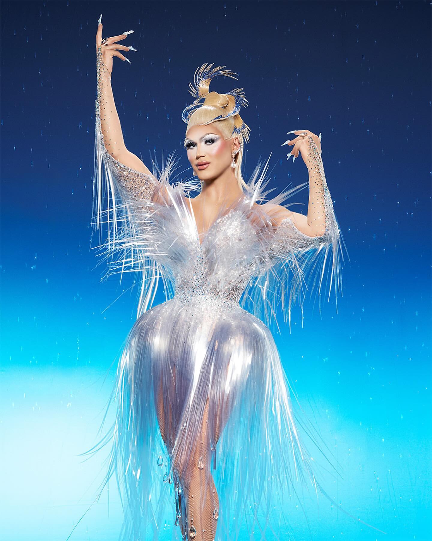 Drag queen poses in a shiny, translucent, tinsel-like dress with long white nails against a gradient light blue background.