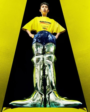 Low-angle shot of an adult wearing a yellow Balenciaga t-shirt with silver metal boot-pants against a yellow and black background.
