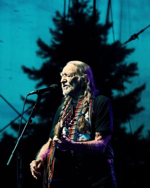 Willie Nelson performs on stage with guitar, singing into a microphone. Silhouette of a tree in blue lighting background.