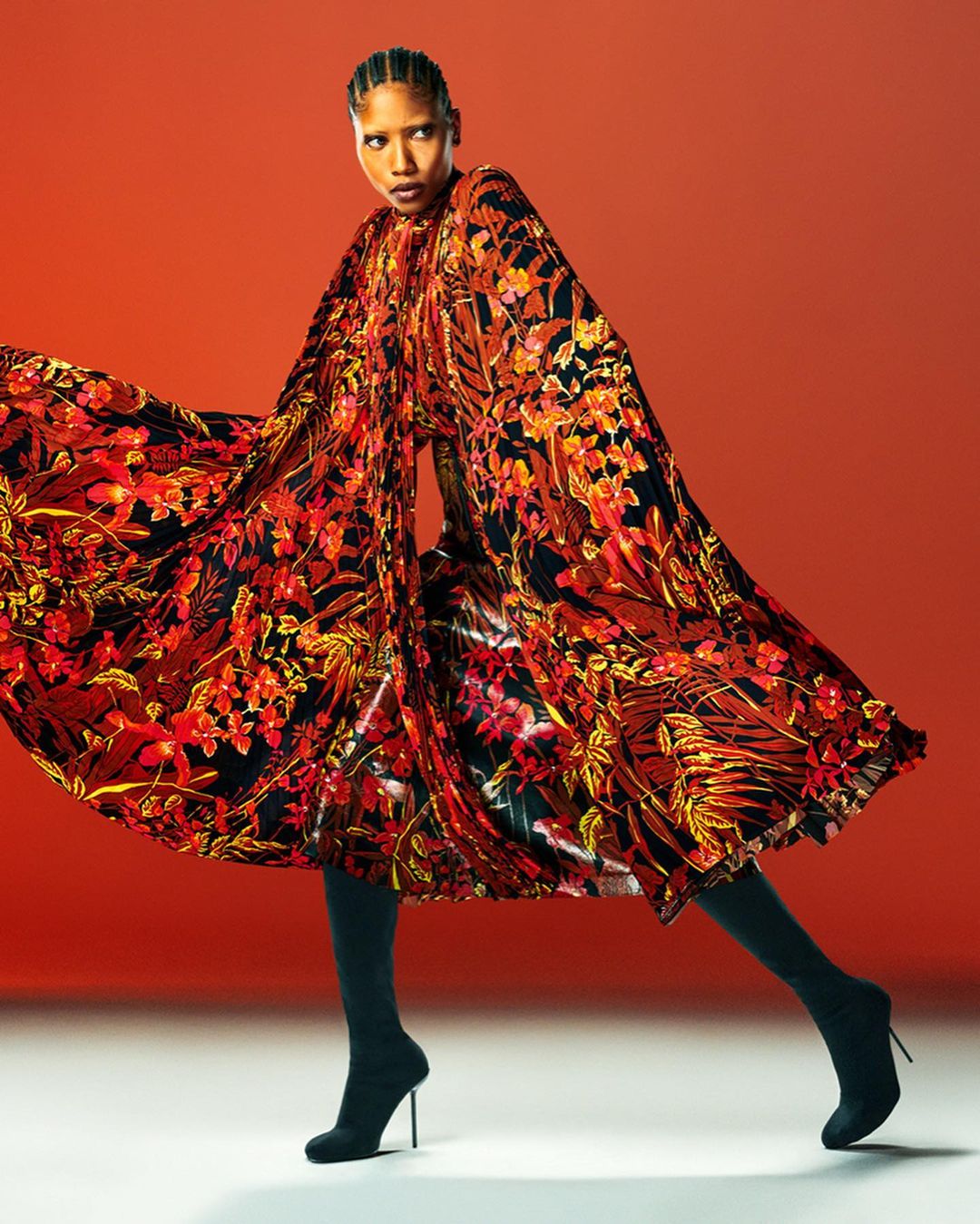 Woman in vibrant floral dress poses against a red background with neutral floor, showcasing bold fashion and dynamic styling.