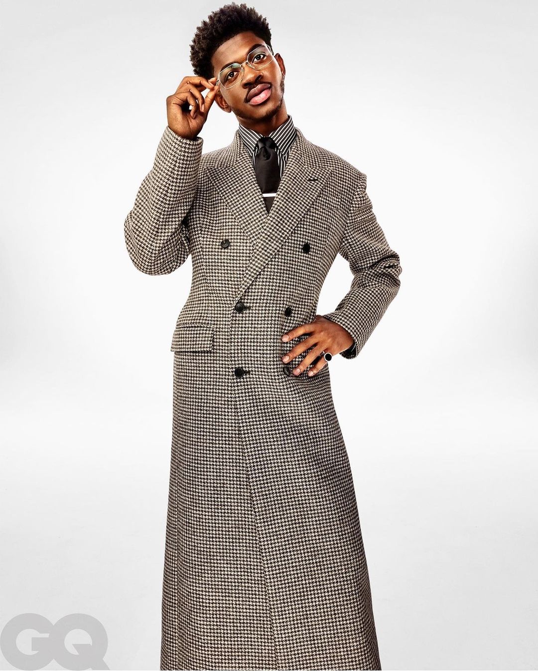 Portrait of African-American male in a houndstooth overcoat and glasses standing in a photo studio against a gradient white backdrop.