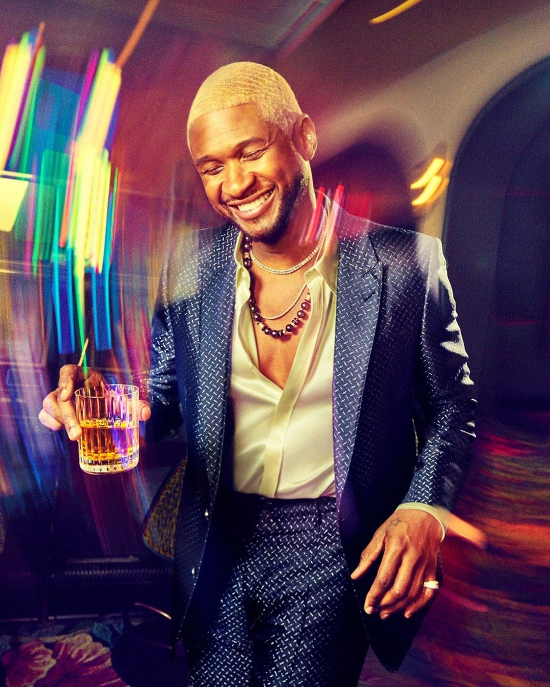Smiling adult male with a drink, wearing a patterned blue suit and jewelry, against a colorful blurred background inside a building.