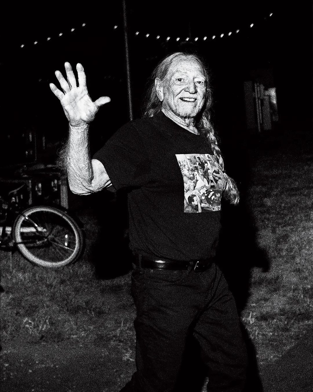 Monochrome portrait of an adult male with long hair, smiling and waving in an outdoor setting with a bicycle visible in the background.