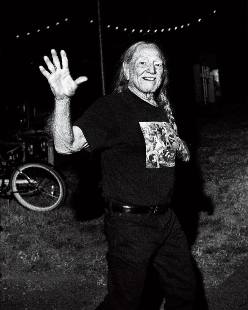 Monochrome portrait of an adult male with long hair, smiling and waving in an outdoor setting with a bicycle visible in the background.