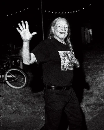Monochrome portrait of an adult male with long hair, smiling and waving in an outdoor setting with a bicycle visible in the background.