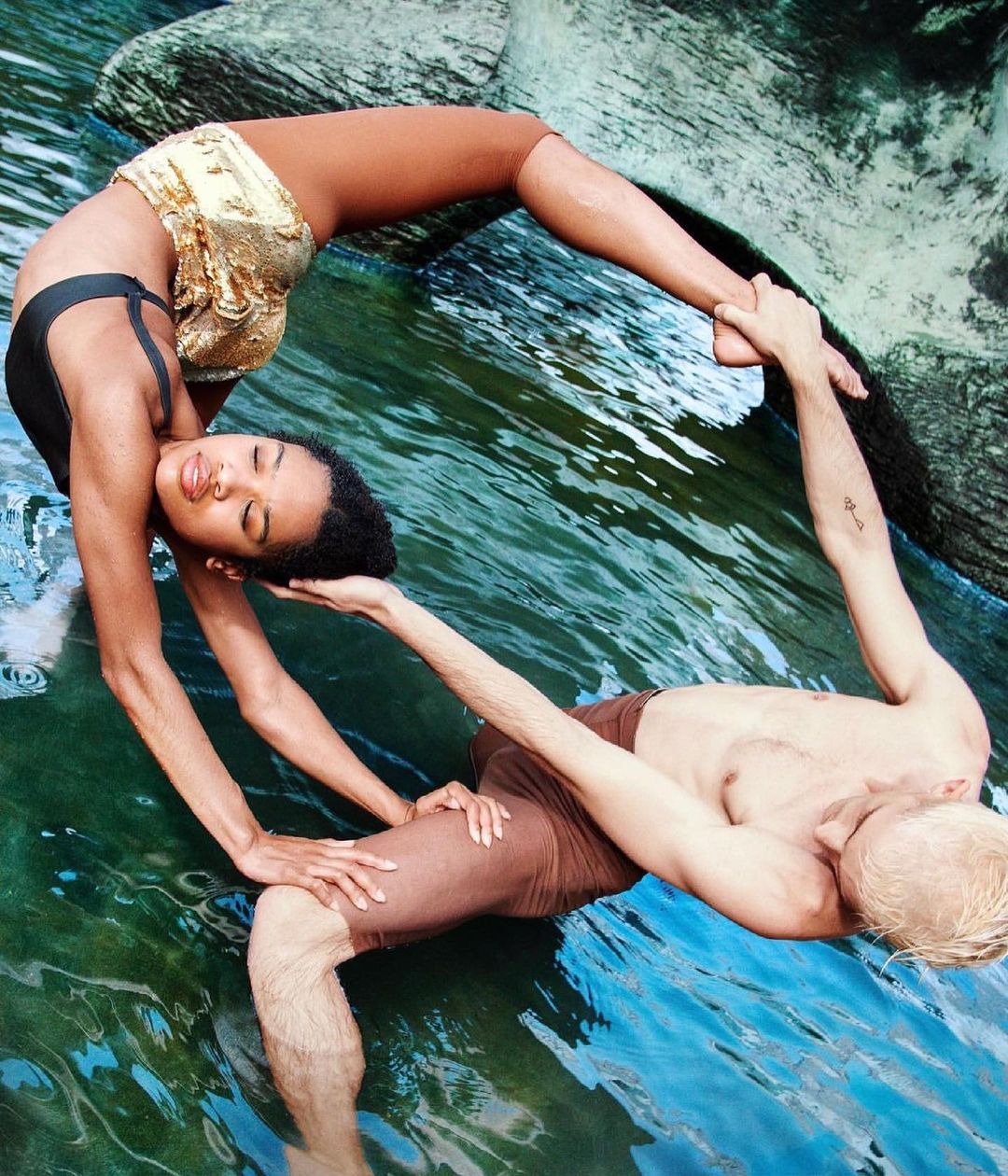 Two people, a woman and a man, performing a yoga-like pose in shallow water with rocky background, captured in a medium shot, showcasing athleticism.