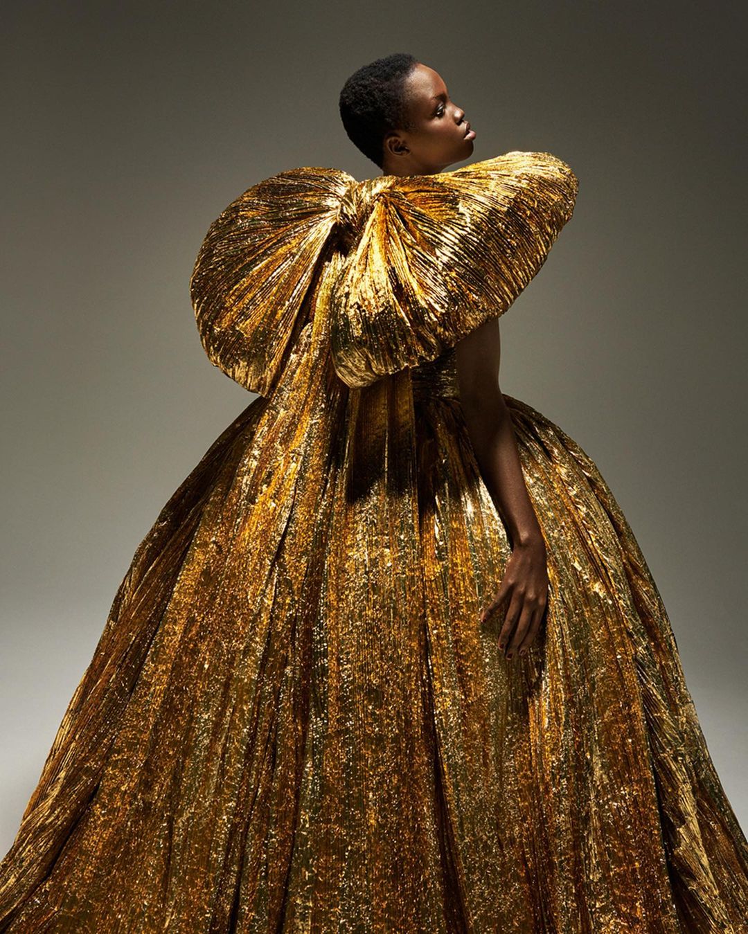 Elegant woman in a dramatic gold gown with a large bow detail, posing in a studio with soft lighting for a fashion editorial shot.