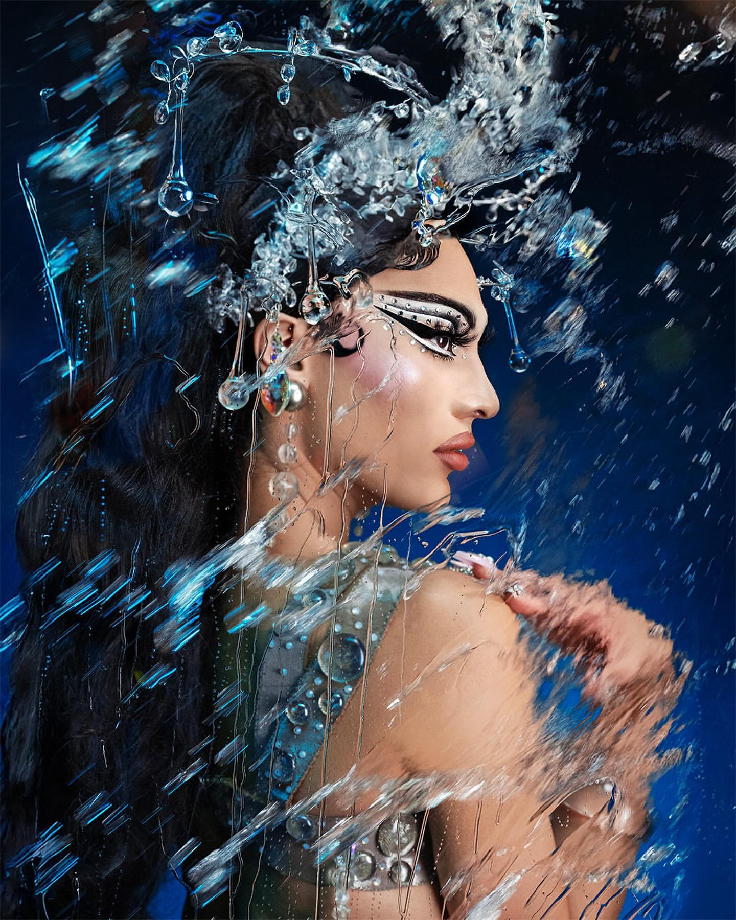 Profile portrait of a model with dramatic eye makeup, adorned with a clear jeweled headpiece and matching earrings, set against a deep blue backdrop.
