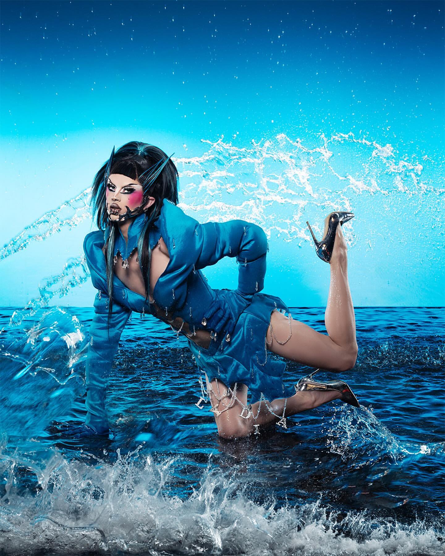 Drag queen posing in a blue outfit and heels, kneeling in splashing water against a blue backdrop with water droplets.